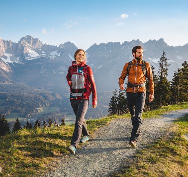 Zwei Personen wandern fröhlich auf einem Bergweg im Hintergrund Gebirgszug.